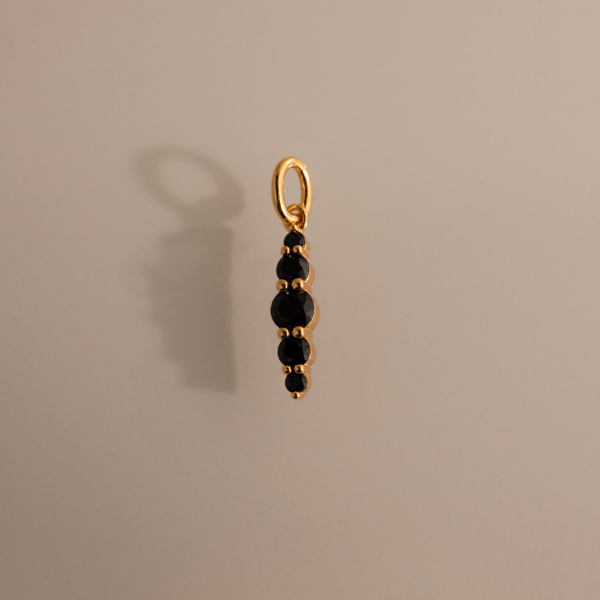 The Ombre Lace Onyx Charm is a gold pendant featuring five small black stones in a vertical line, casting an elegant shadow on a beige surface.