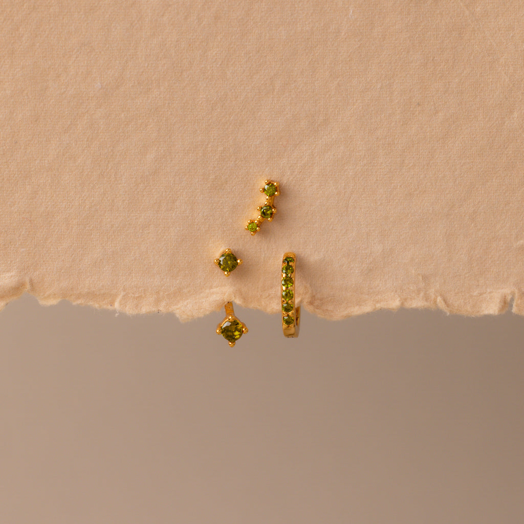The Peridot Earring Trio Set, featuring four gold pieces with green stones including a pavé huggie hoop, is displayed on textured, torn beige paper.