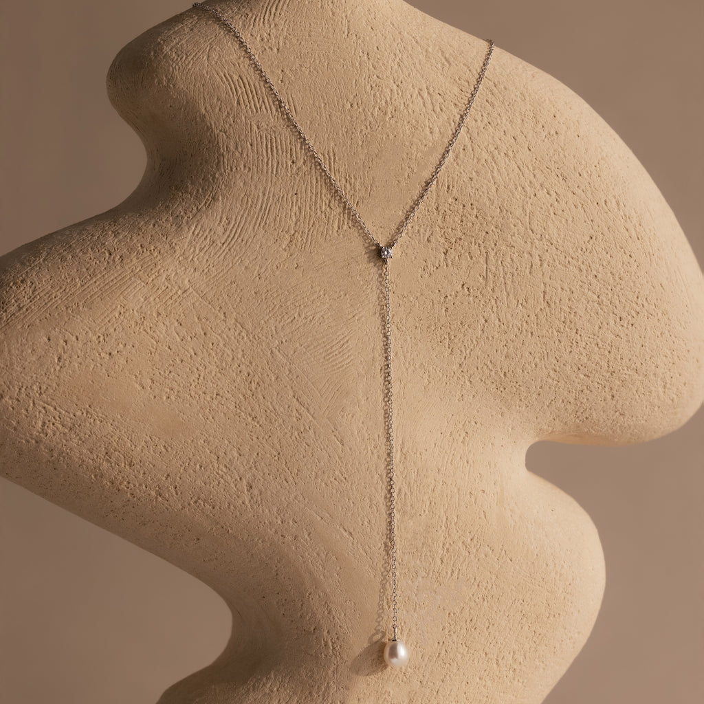 The Pearl Drop Lariat Necklace in Sterling Silver, featuring a delicate vertical chain and pearl pendant, drapes elegantly over a textured abstract beige sculpture—a stunning piece of elegant jewelry.