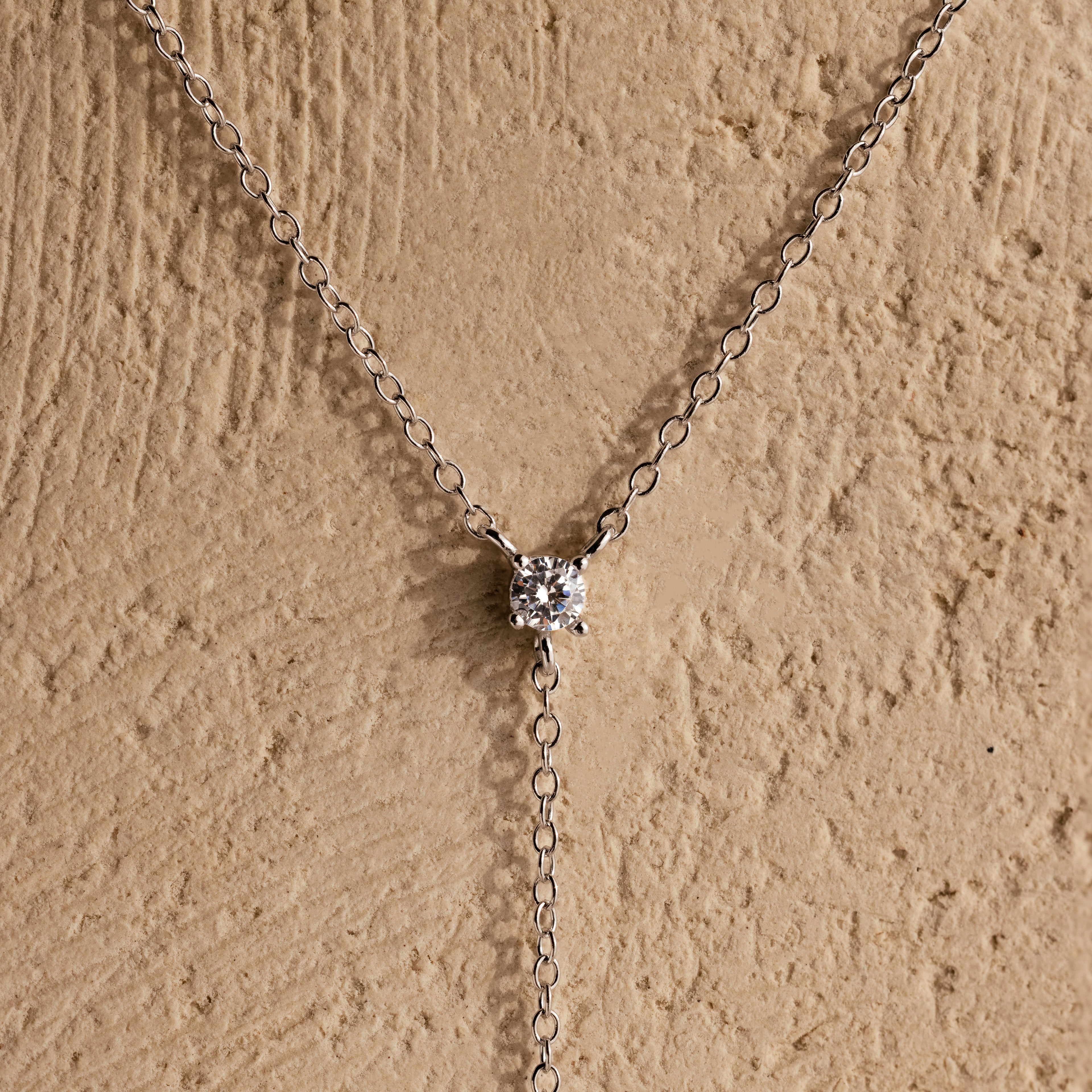 The Pearl Drop Lariat Necklace in Sterling Silver features a delicate vertical chain with a single pearl pendant, elegantly displayed against a textured beige background—an ideal choice for lovers of refined jewelry.