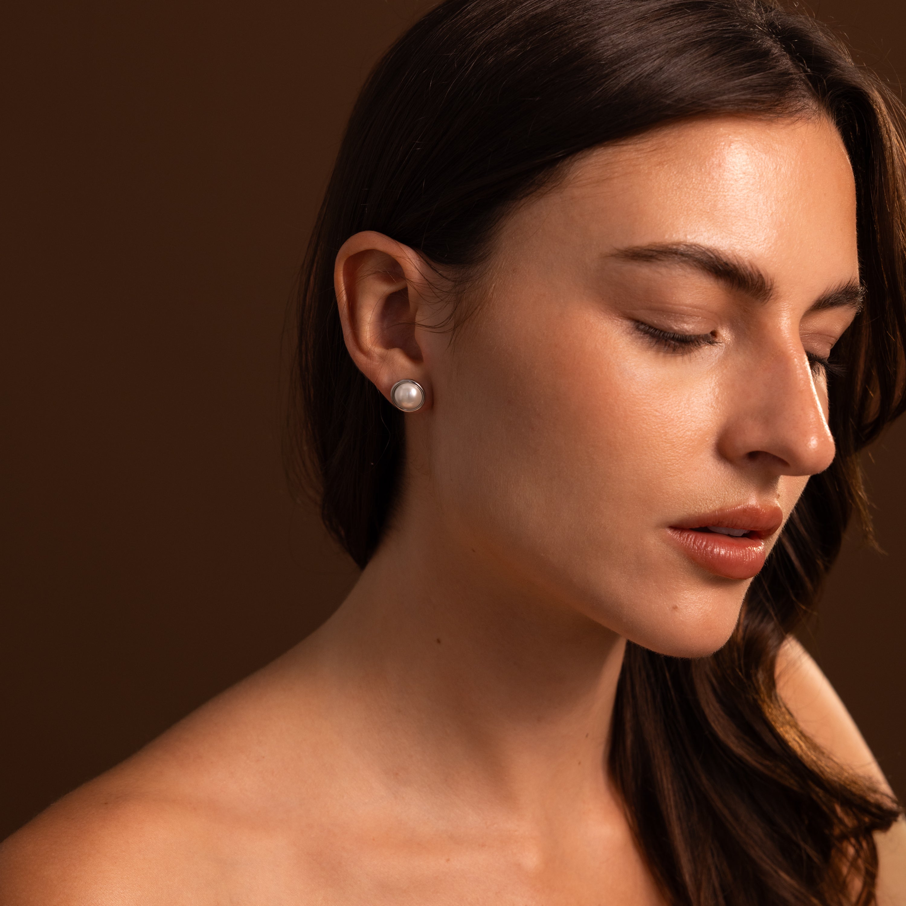 Woman with long brown hair wears Pearl Bezel Studs in Sterling Silver, looking down against a brown background.