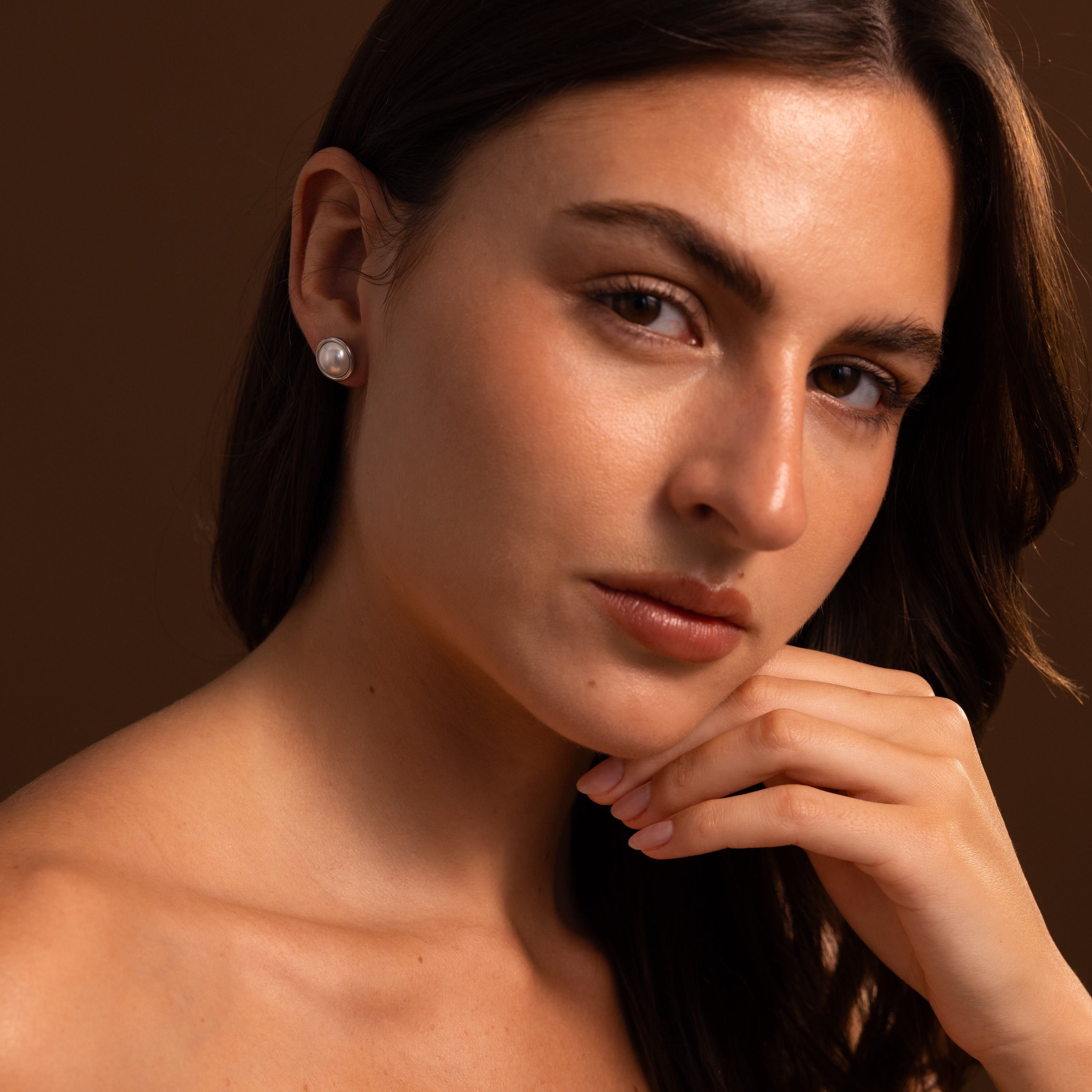 Woman with long brown hair wears Pearl Bezel Studs in Sterling Silver, posing with her hand under her chin against a brown background.