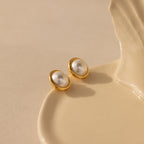 The Pearl Bezel Studs in 18K Gold rest elegantly on a cream ceramic dish, their luminous pearls and gold rims softly shining against a beige background—ideal for everyday wear.