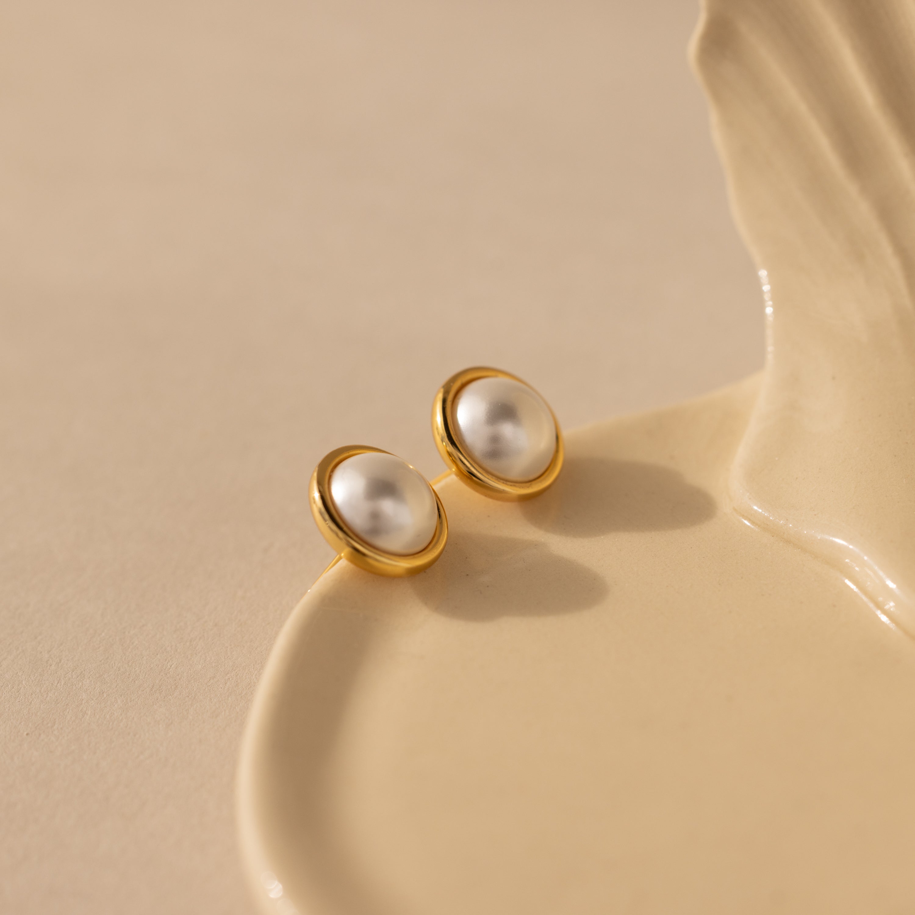 The Pearl Bezel Studs in 18K Gold rest elegantly on a cream ceramic dish, their luminous pearls and gold rims softly shining against a beige background—ideal for everyday wear.