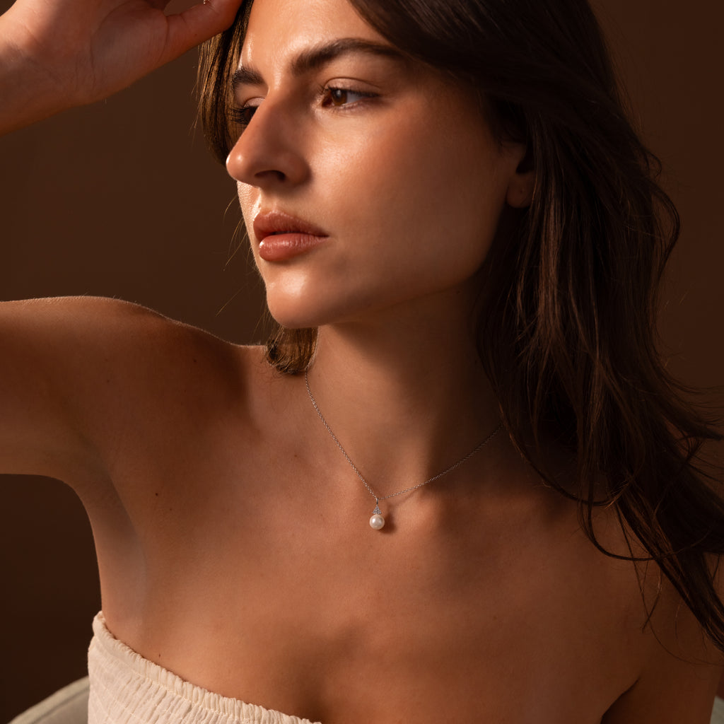 A woman in a strapless top gazes sideways, highlighting the Diamond Halo Pearl Necklace in Sterling Silver—a timeless piece—set against a brown background.