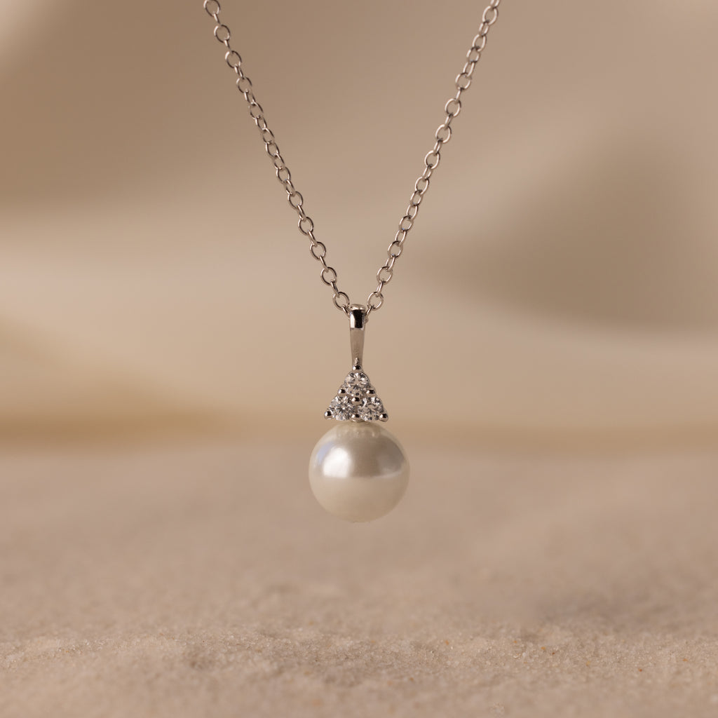 The Diamond Halo Pearl Necklace showcases a round pearl pendant encircled by sparkling diamonds, elegantly displayed on a soft, sandy background—a timeless and exquisite jewelry piece.