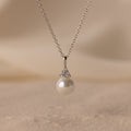 The Diamond Halo Pearl Necklace showcases a round pearl pendant encircled by sparkling diamonds, elegantly displayed on a soft, sandy background—a timeless and exquisite jewelry piece.