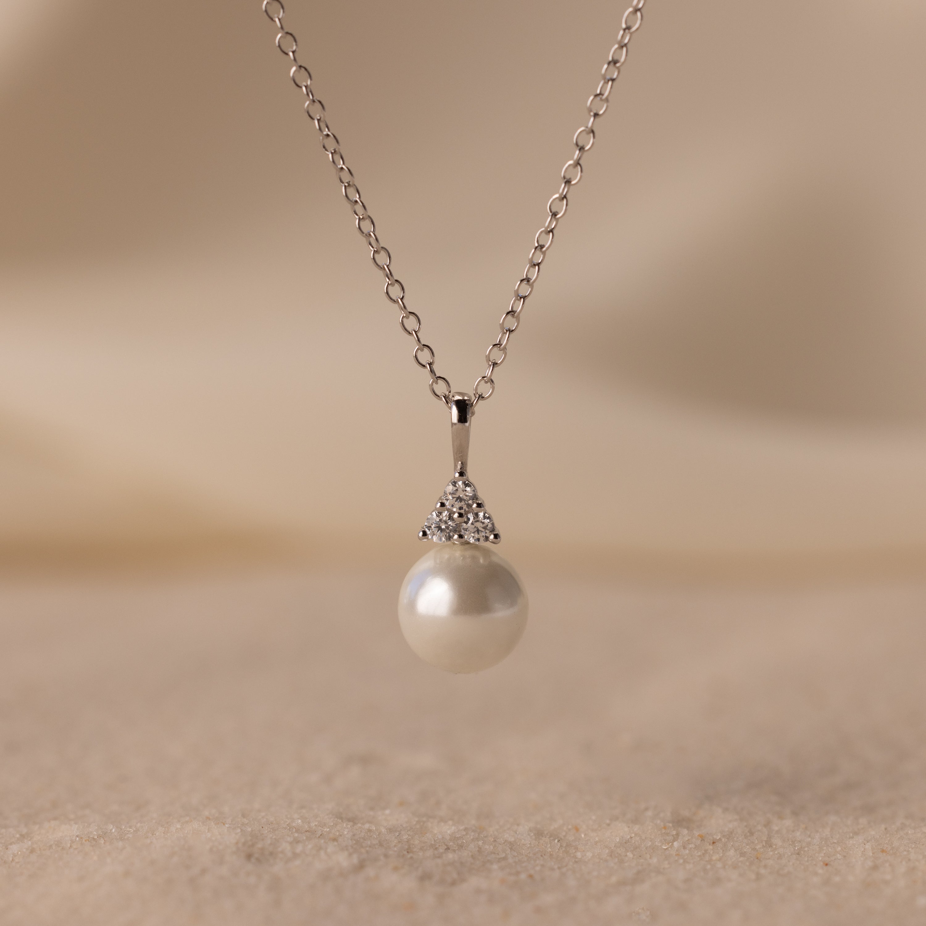 The Diamond Halo Pearl Necklace showcases a round pearl pendant encircled by sparkling diamonds, elegantly displayed on a soft, sandy background—a timeless and exquisite jewelry piece.