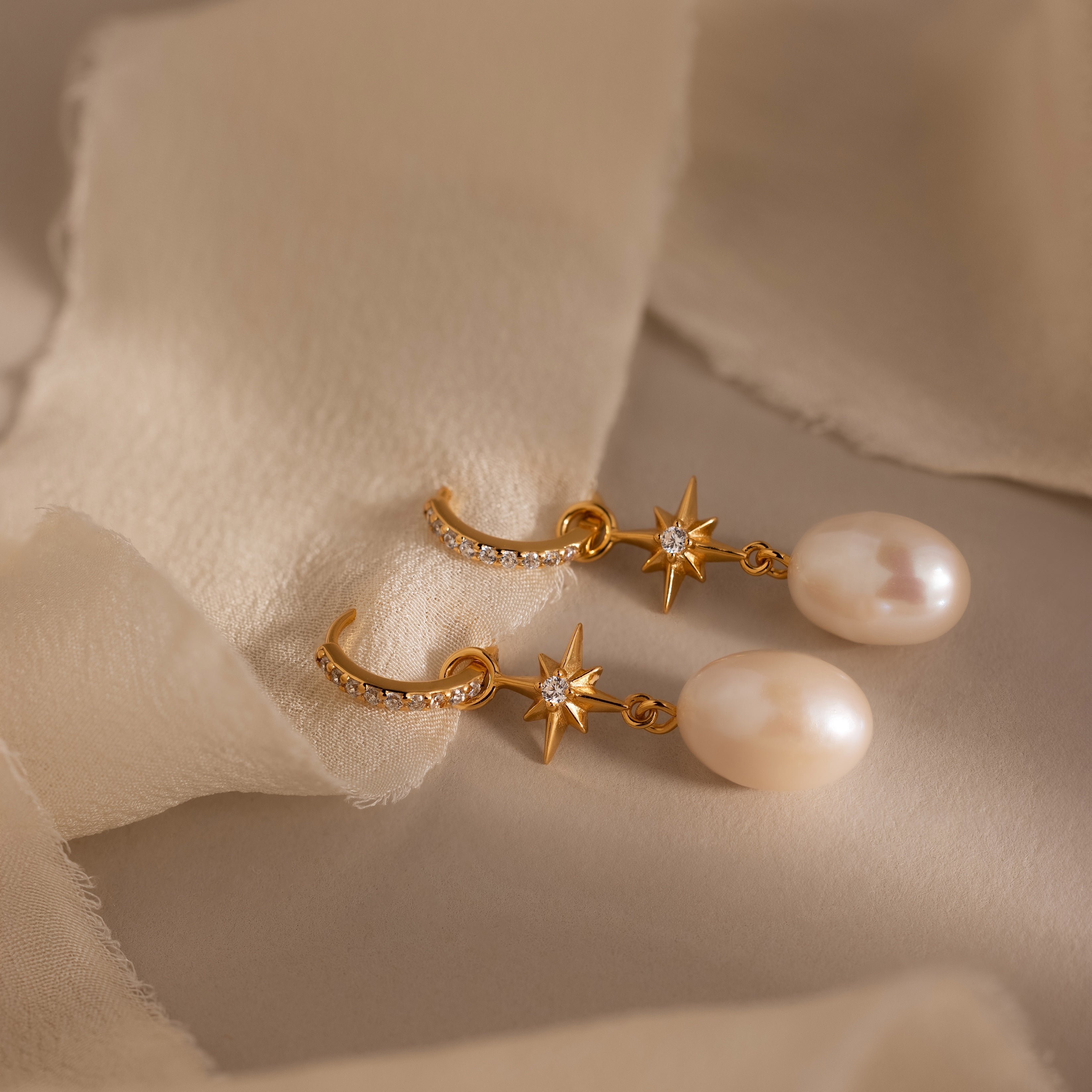 The North Star Pearl Hoops in 18K Gold, featuring star accents, pearls, and small diamonds, are displayed on soft beige fabric—a stunning celestial-inspired jewelry piece.
