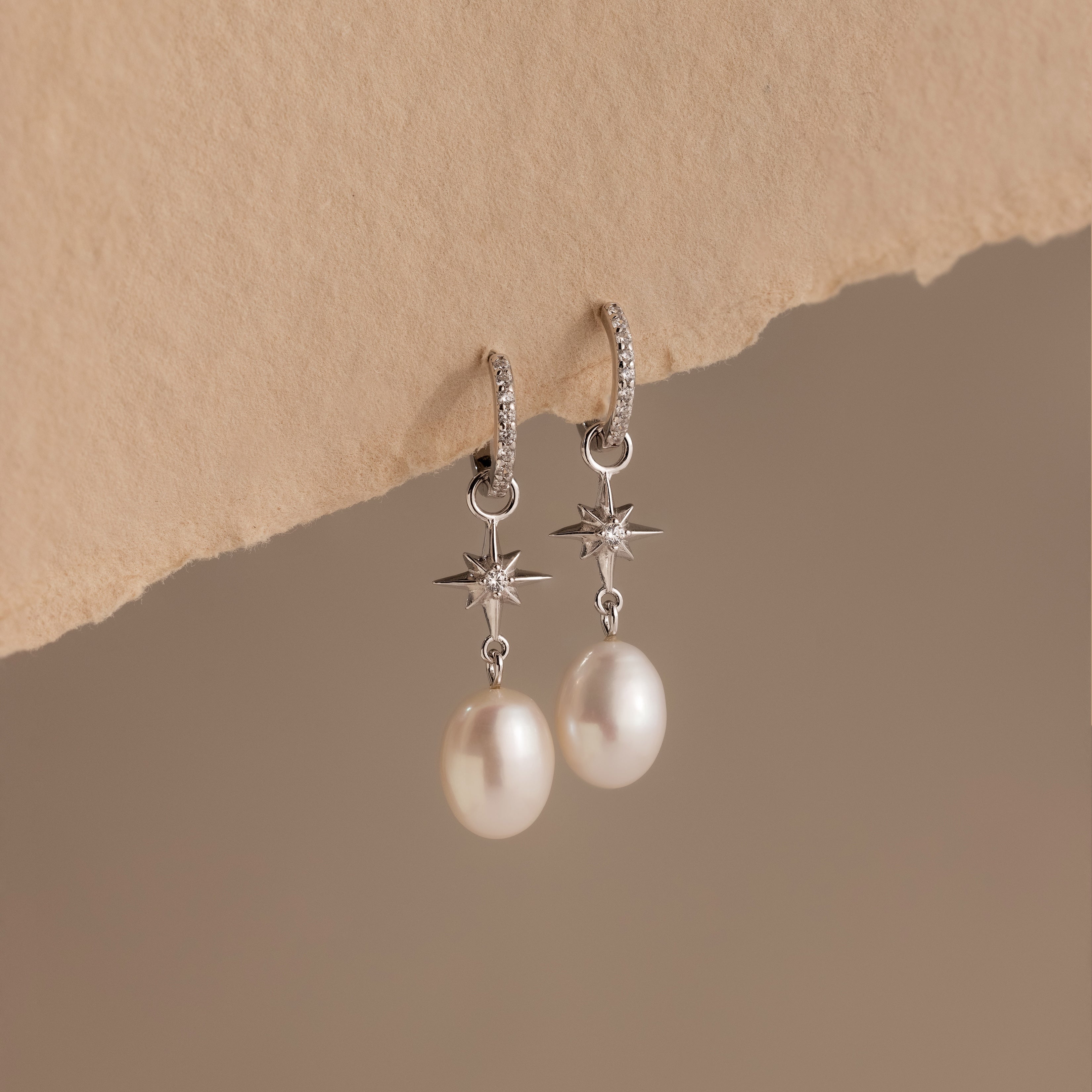 The North Star Pearl Hoops in Sterling Silver, featuring star details and dangling pearls, hang from textured beige paper against a neutral background.