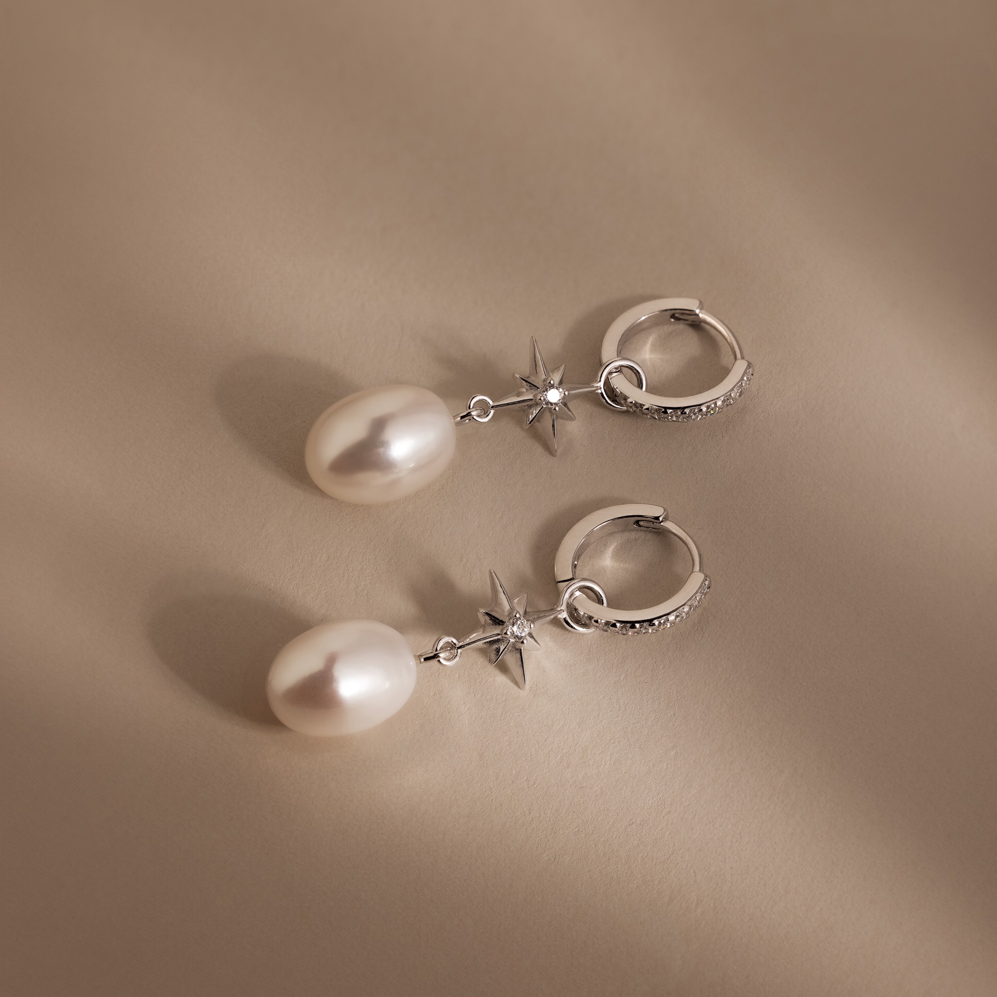 The North Star Pearl Hoops in Sterling Silver feature star accents and dangling pearls for a celestial look, shown on a beige background.
