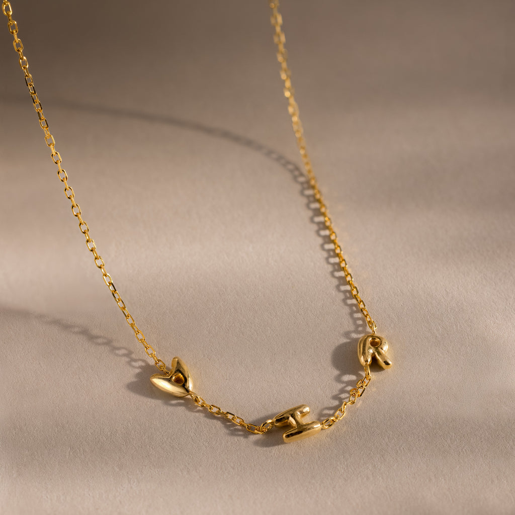 Up close of a gold dainty bubble initial charm necklace with letters "AHR" resting on a neutral background.