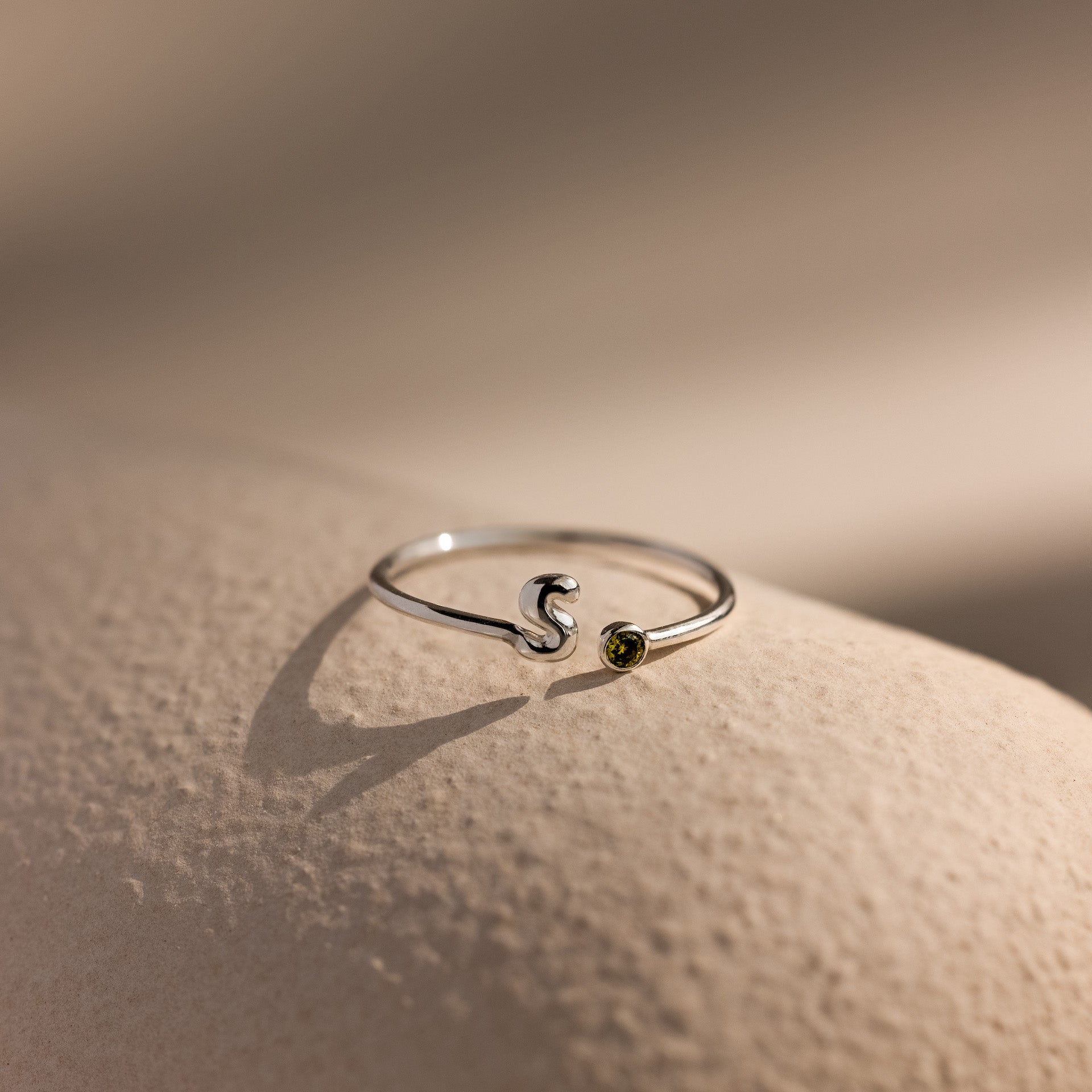 Up close of a silver bubble letter duo gemstone ring with the initial "S" and a green peridot birthstone resting on a neutral surface.