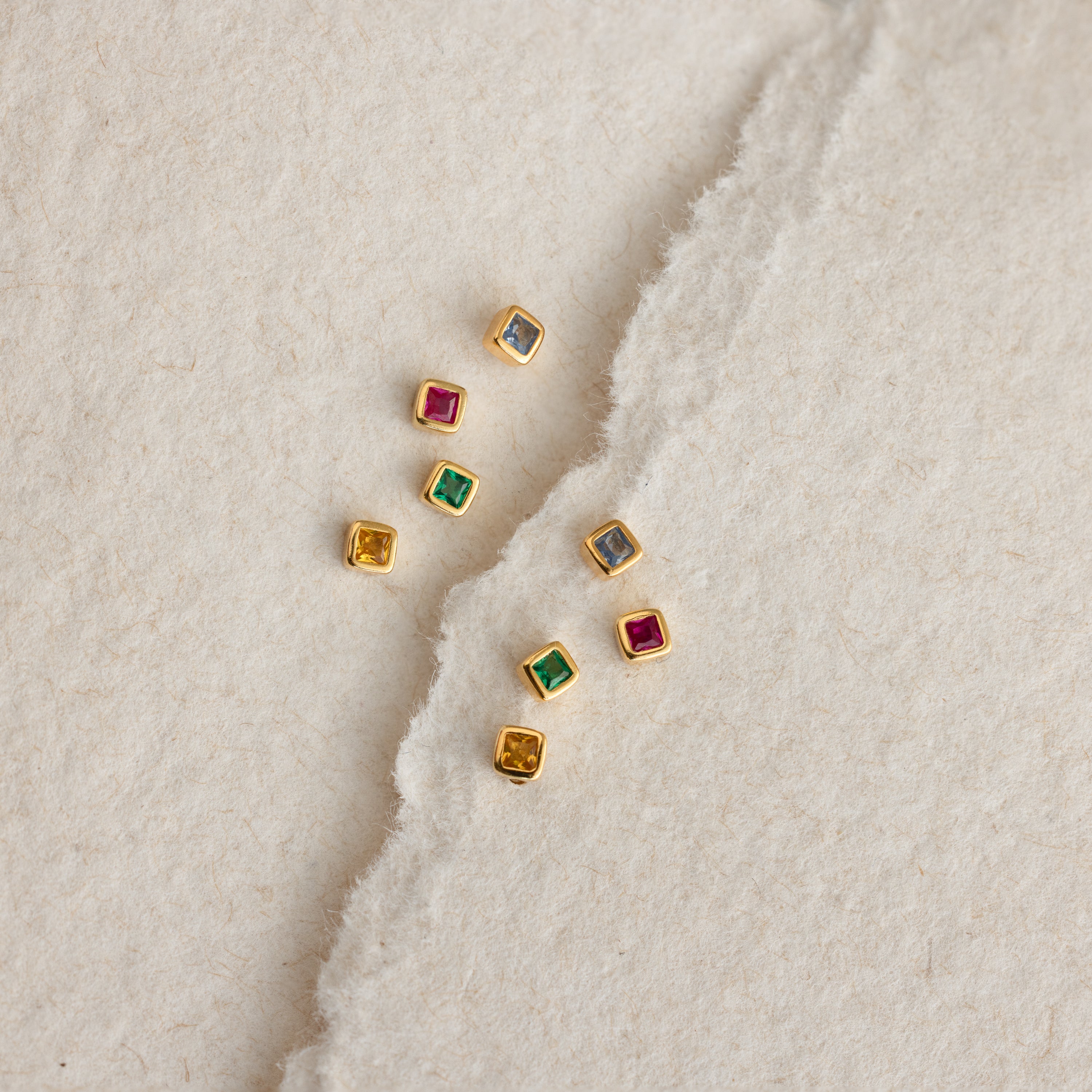 Eight small Square Bezel Birthstone Flatback Studs, each with a colored gemstone, are displayed on textured torn cream paper.