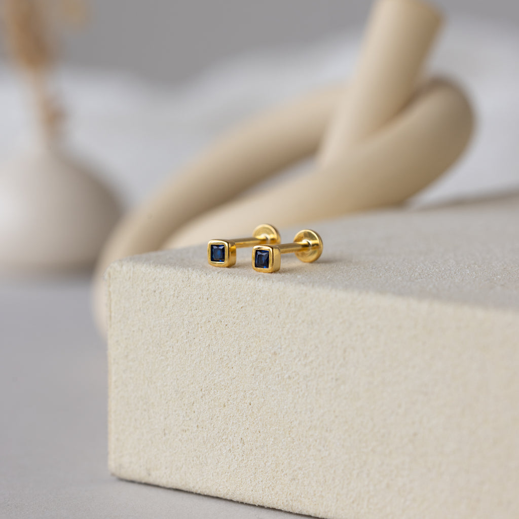 Square Bezel Sapphire Flatback Studs showcasing small sapphire CZ gemstones in gold, elegantly presented on a cream-colored textured surface.