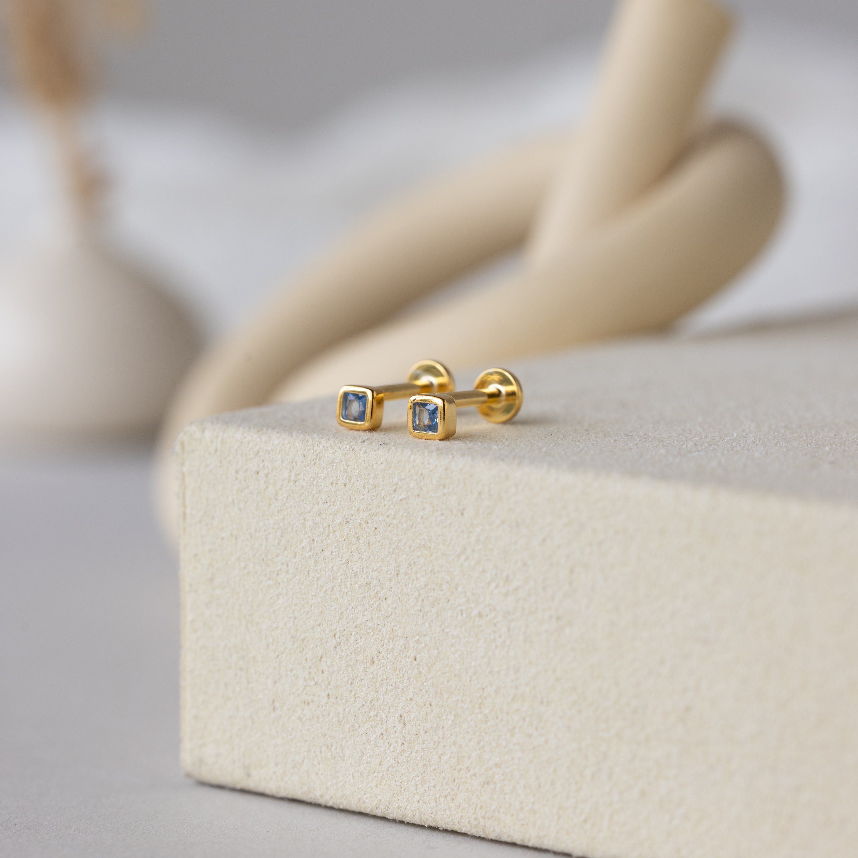 A pair of Square Bezel Blue Zircon Flatback Studs with square-cut CZ gemstones is displayed on a textured beige surface.