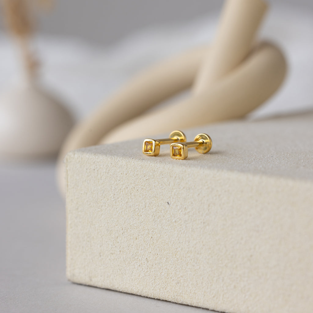 Square Bezel Topaz Flatback Studs—small gold earrings with square topaz accents—are displayed on a beige textured surface with a blurred background.