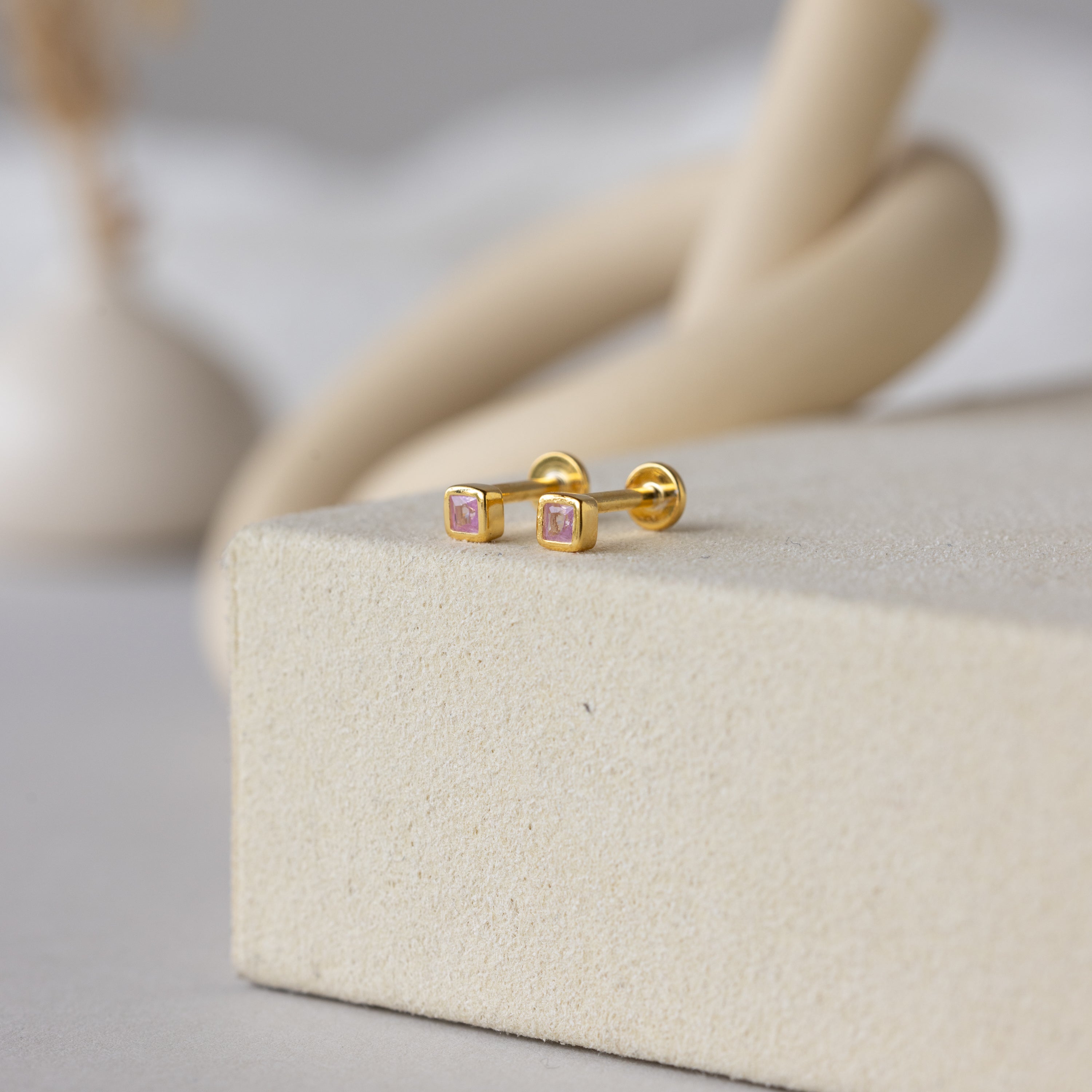 Square Bezel Pink Tourmaline Flatback Studs with gold finish and pink gemstones rest on a beige textured surface, bringing an elegant touch to any look.