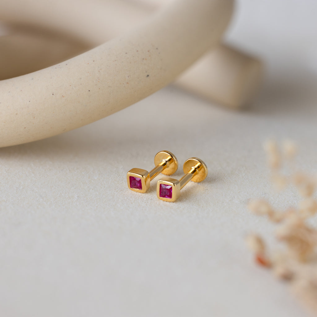 Square Bezel Ruby Flatback Studs, featuring small CZ gemstones set in a gold square bezel, are displayed on a light, textured surface with soft decor.