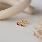 Square Bezel Ruby Flatback Studs, featuring small CZ gemstones set in a gold square bezel, are displayed on a light, textured surface with soft decor.