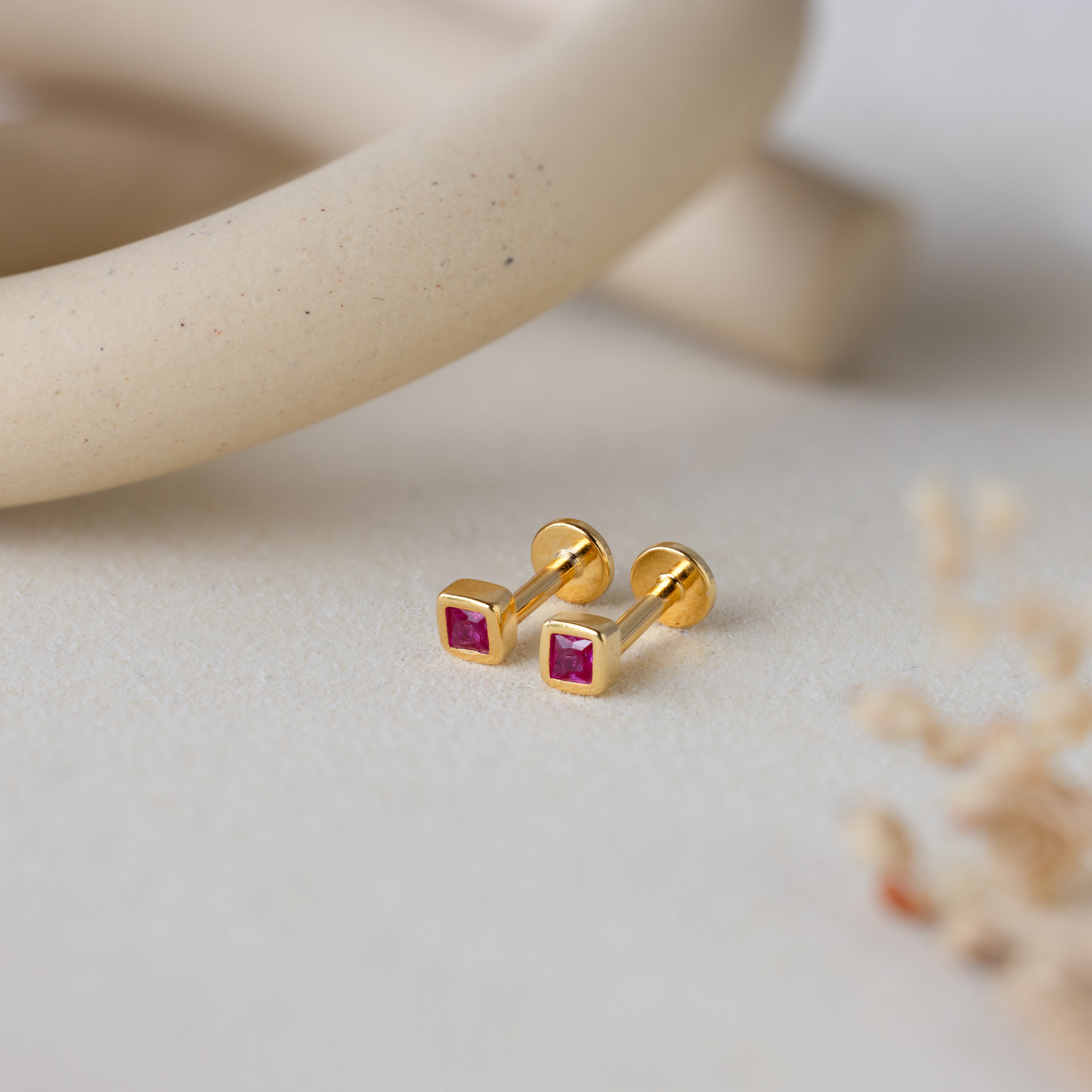 Square Bezel Ruby Flatback Studs, featuring small CZ gemstones set in a gold square bezel, are displayed on a light, textured surface with soft decor.