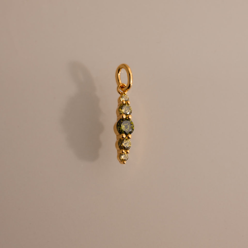 The Ombre Lace Peridot Charm is a gold pendant featuring four small gemstones arranged vertically, adding a personalized touch against a neutral background.