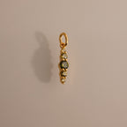 The Ombre Lace Peridot Charm is a gold pendant featuring four small gemstones arranged vertically, adding a personalized touch against a neutral background.