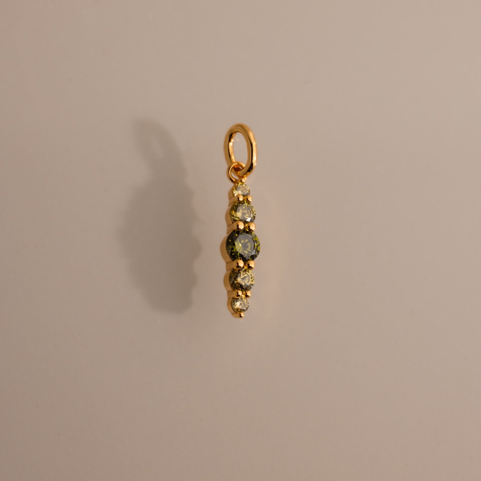 The Ombre Lace Peridot Charm is a gold pendant featuring four small gemstones arranged vertically, adding a personalized touch against a neutral background.