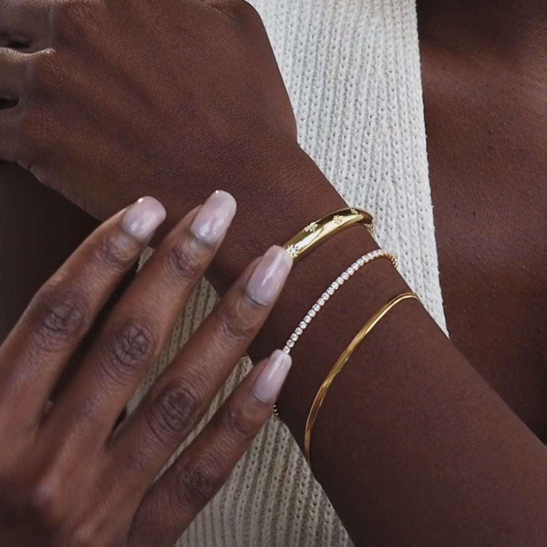 Up close of model wearing a stacked set of a gold CZ diamond star bangle bracelet, diamond tennis bracelet, and gold snake chain bracelet.
