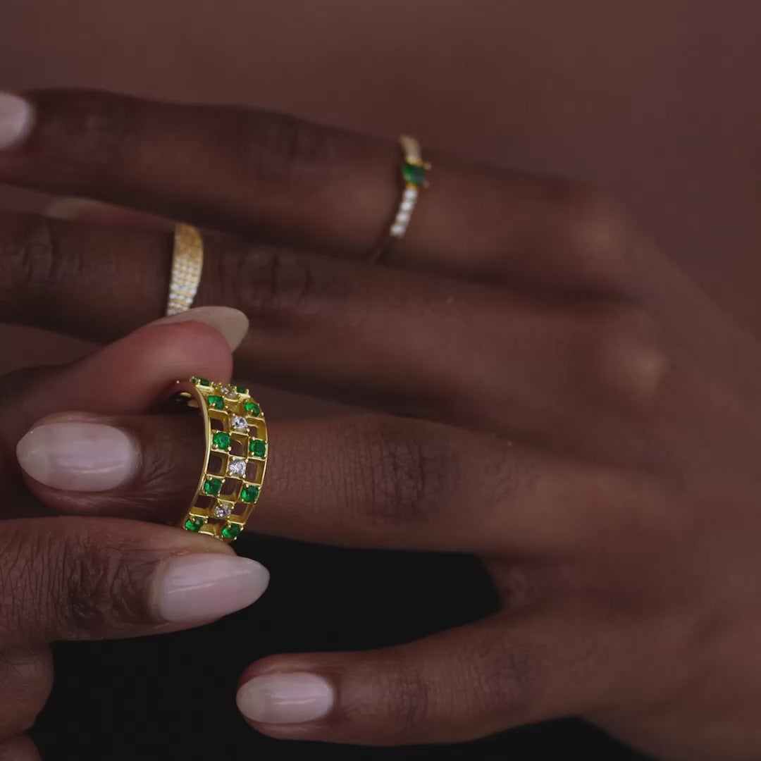 Model showcasing a gold checkered band ring featuring alternating rows of emerald and diamond round-cut CZ stones set in an openwork grid design, stacked with multiple gold rings.