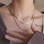 Model wearing a gold dainty bubble K letter charm necklace, layered with a gold herringbone necklace and gold box chain necklace.