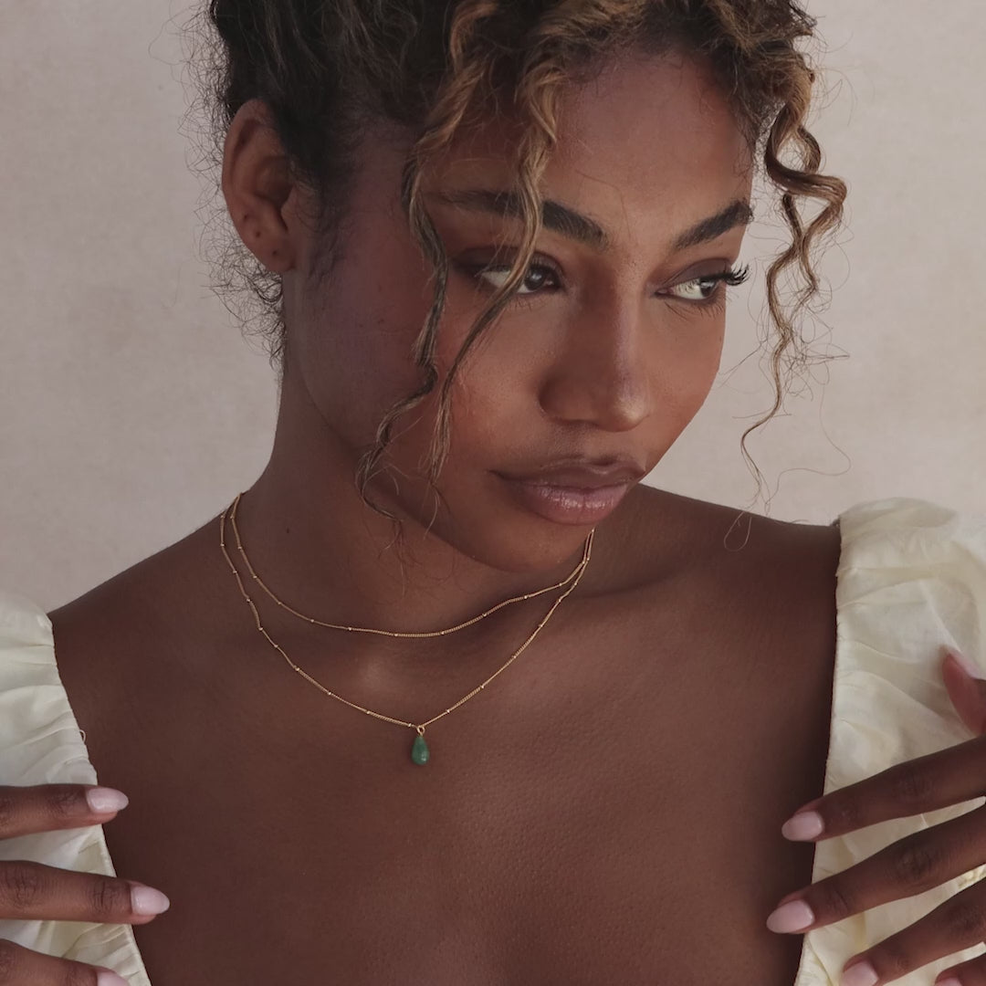 Model showcasing a gold satellite chain necklace featuring a single green jade teardrop gemstone pendant, layered with a matching gold satellite necklace.