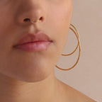 Model showcasing two large, textured gold hoop earrings. The hammered finish on the hoops reflects light, creating a shimmering effect.