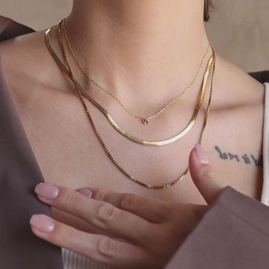 Model wearing a gold dainty bubble K letter charm necklace, layered with a gold herringbone necklace and gold box chain necklace.
