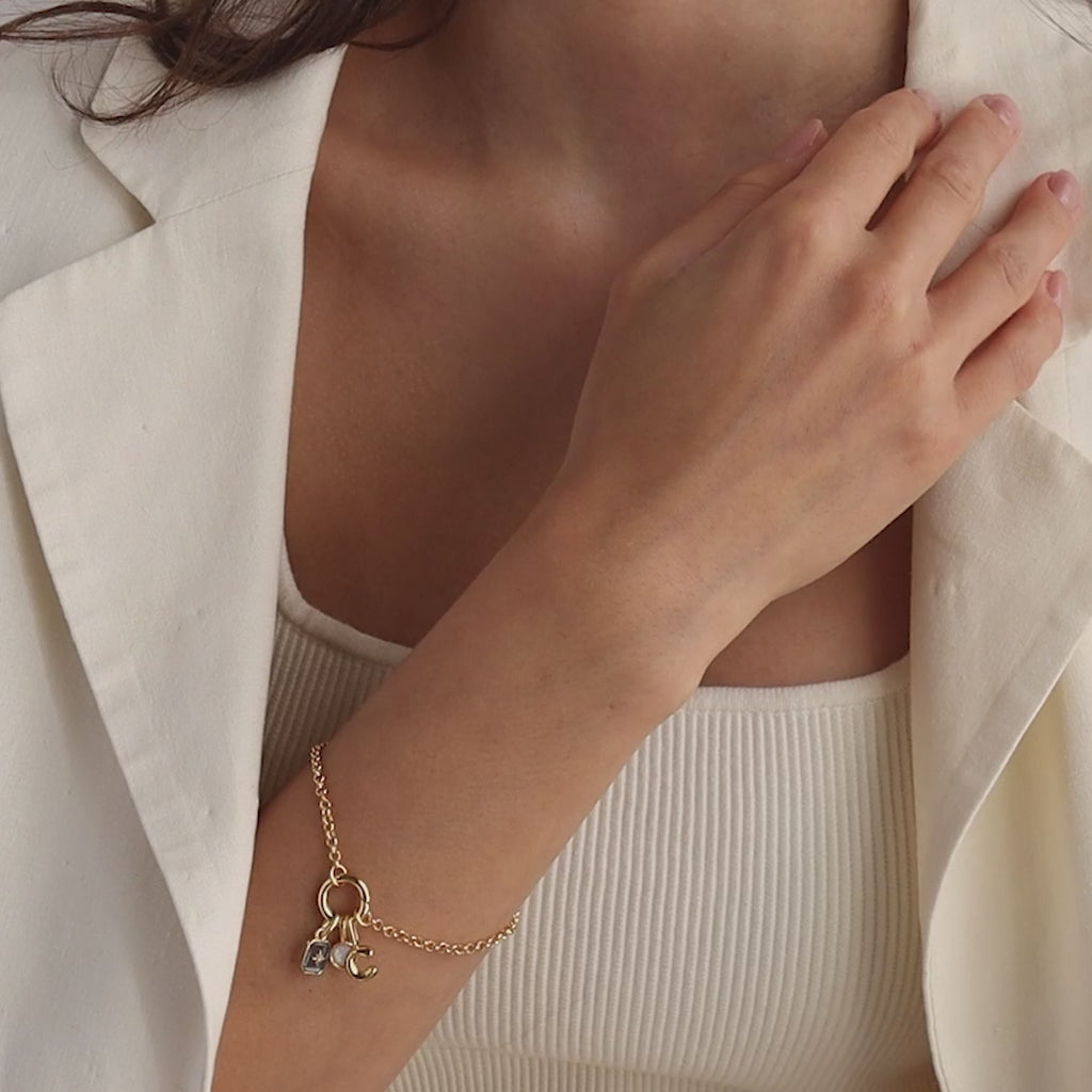 Model wearing a gold rolo charm bracelet with a blue aquamarine north star charm, bubble letter "C" charm, and an opal signet charm.