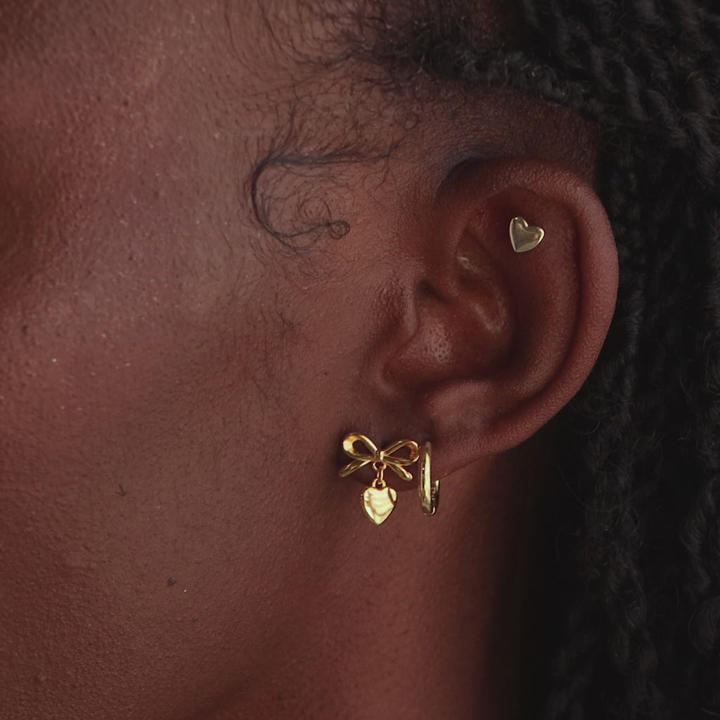 Model wearing the gold bow heart stud earring paired with a small gold heart cartilage stud and a hoop earring.