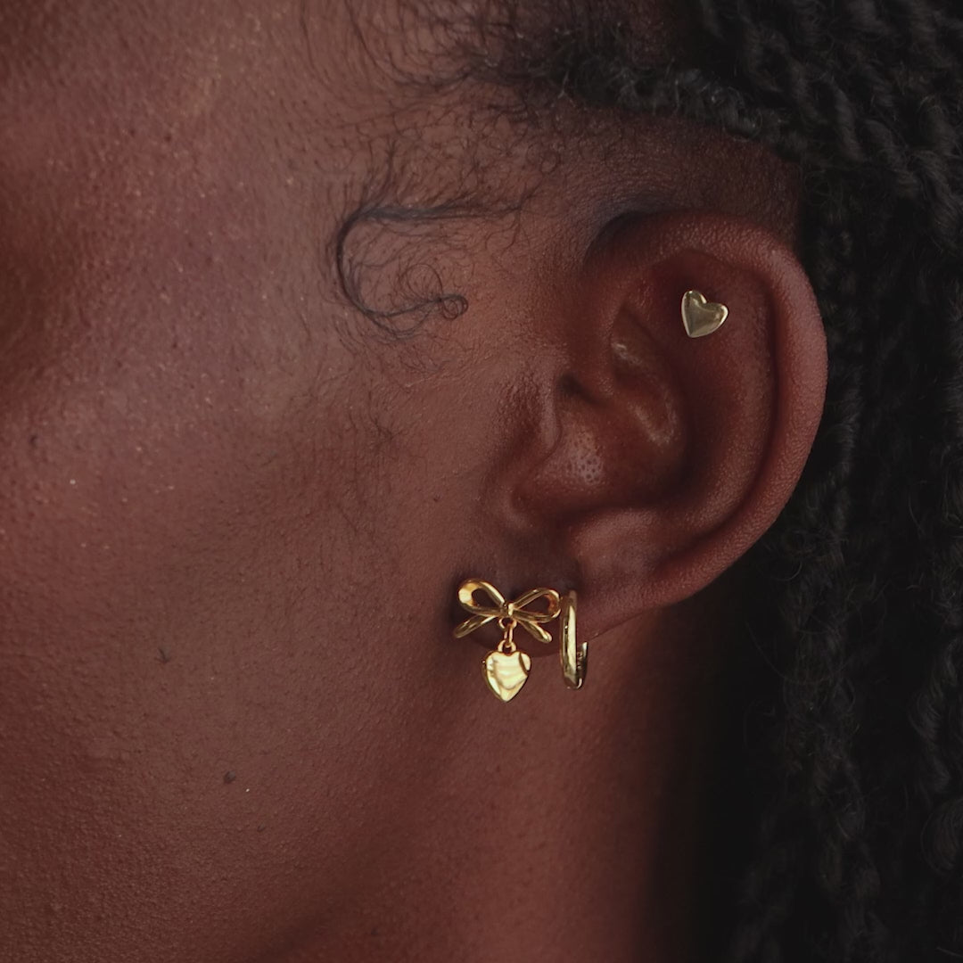 Model wearing the gold bow heart stud earring paired with a small gold heart cartilage stud and a hoop earring.