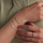 Model wearing a dainty gold bubble letter bracelet of the name "ALEXA" with a sapphire birthstone, stacked with a snake chain bracelet and pave heart bracelet.
