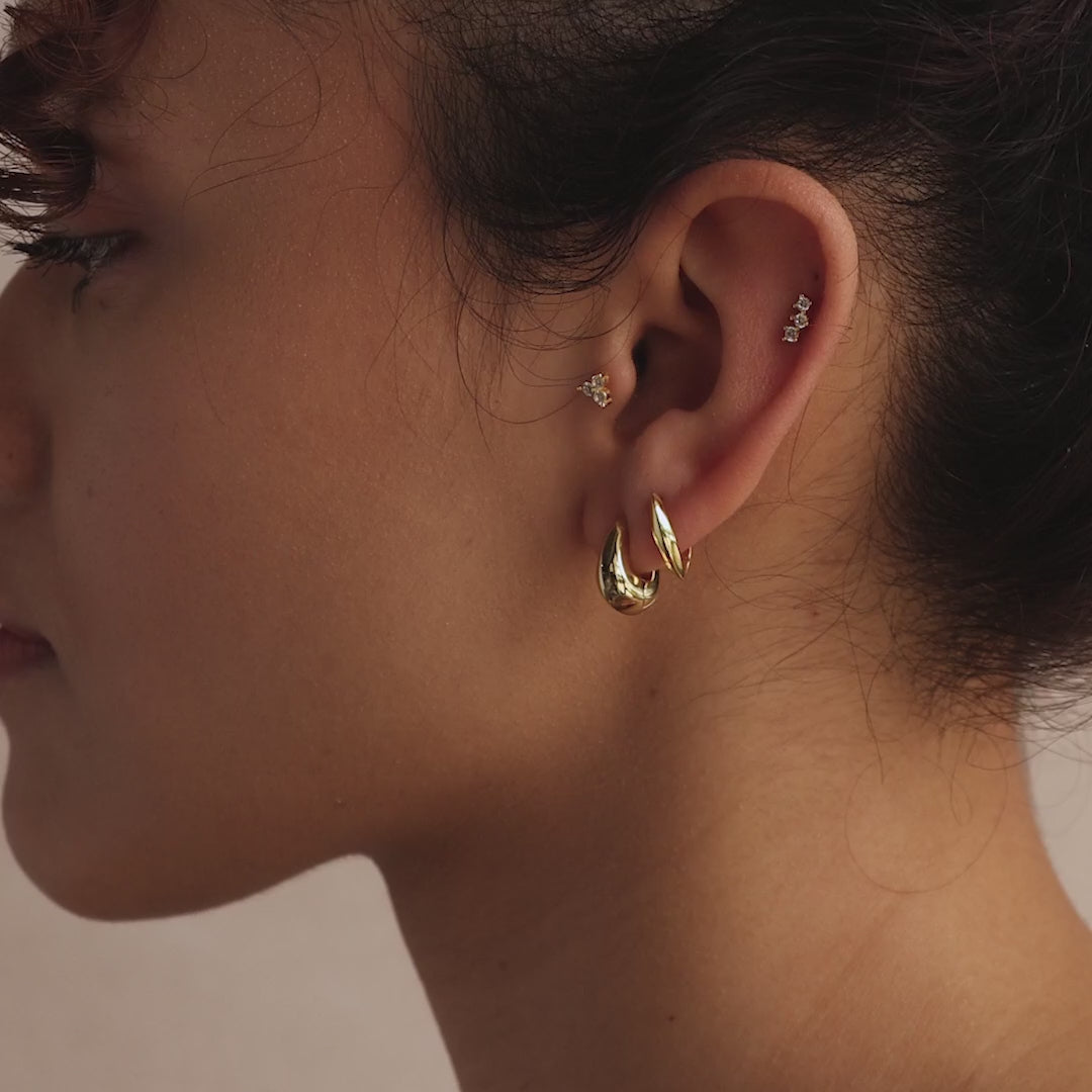 Close-up of a model’s ear adorned with multiple gold earrings, including a bold leaf-shaped hoop and a smaller pointed hoop, paired with delicate gemstone studs in the upper ear.