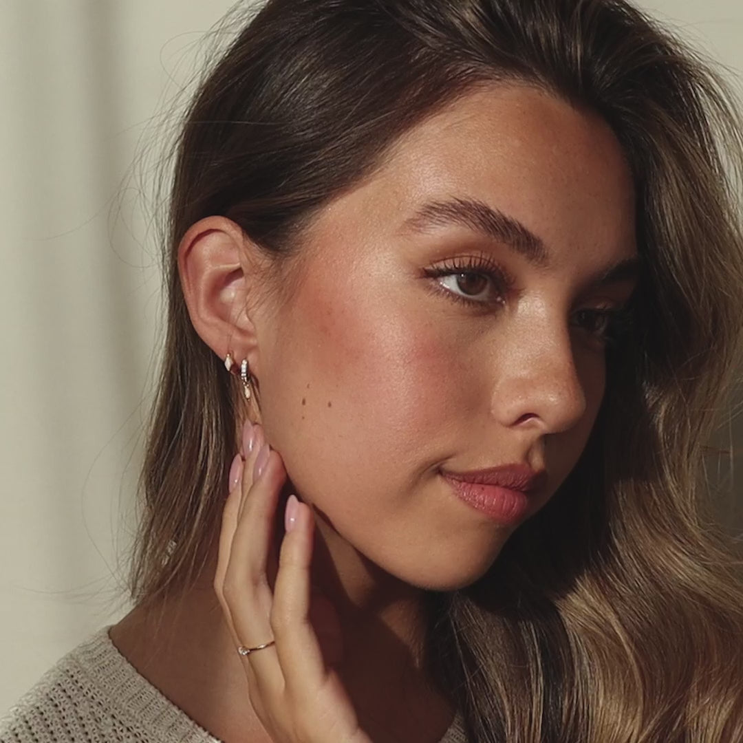 A  woman wearing multiple earrings on her ear, including a gold huggie with a small rhombus-shaped gemstone among other minimal hoops, with soft glam makeup and natural lighting.