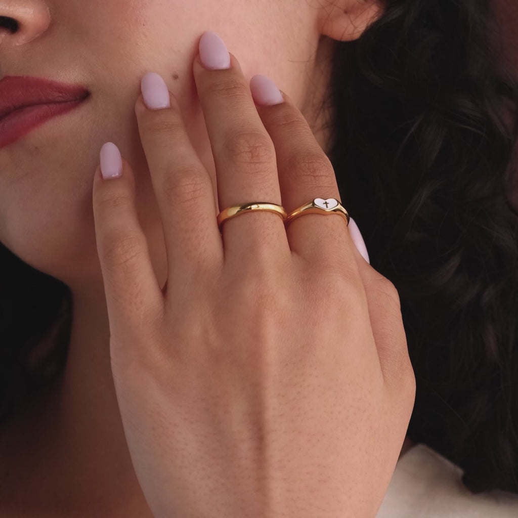 Model a gold heart-shaped ring with a white enamel face and small gold cross, styled with a matching gold ring.
