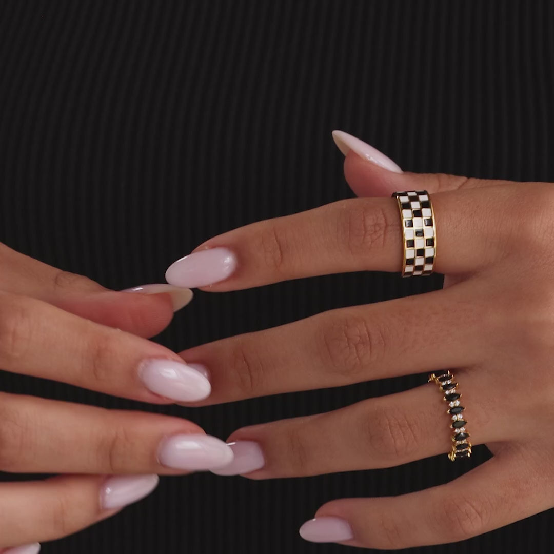 Model showcasing a thick gold band ring with a black and white enamel checkerboard pattern, stacking with multiple gold rings with black onyx gemstones.
