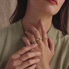 Model wearing a dainty gold bubble letter birthstone duo ring with the initial "A" and a clear diamond gemstone, stacked with multiple gold rings with pink ruby gemstones.