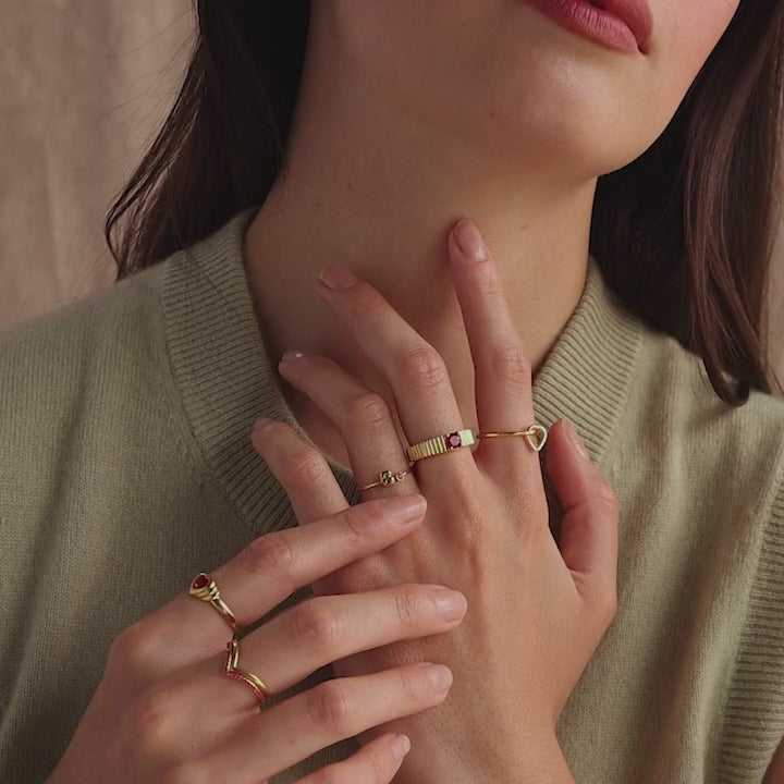 Model wearing a dainty gold bubble letter birthstone duo ring with the initial "A" and a clear diamond gemstone, stacked with multiple gold rings with pink ruby gemstones.