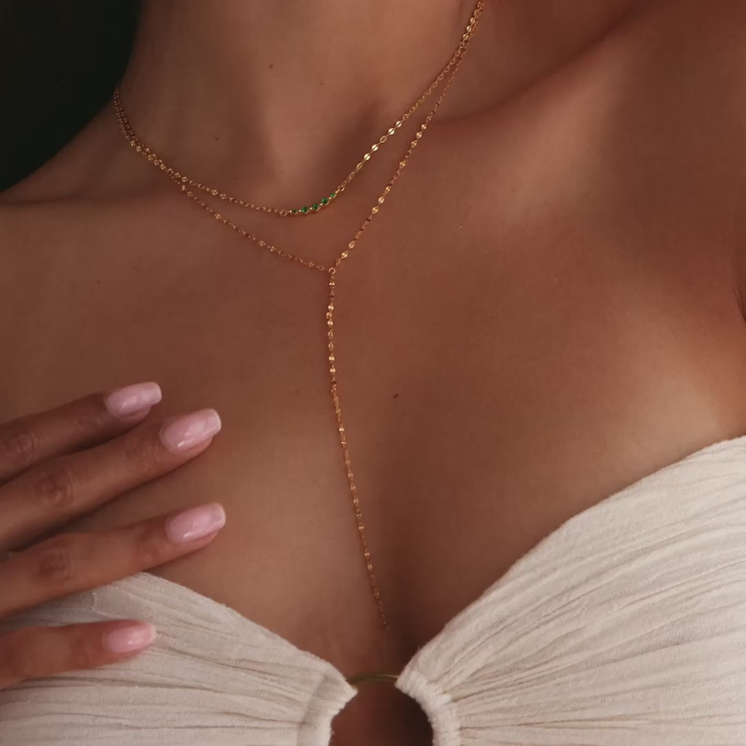 Model showcasing a gold dainty mirror chain lariat necklace, layered with a gold necklace with an arc-shaped charm featuring an emerald birthstone.