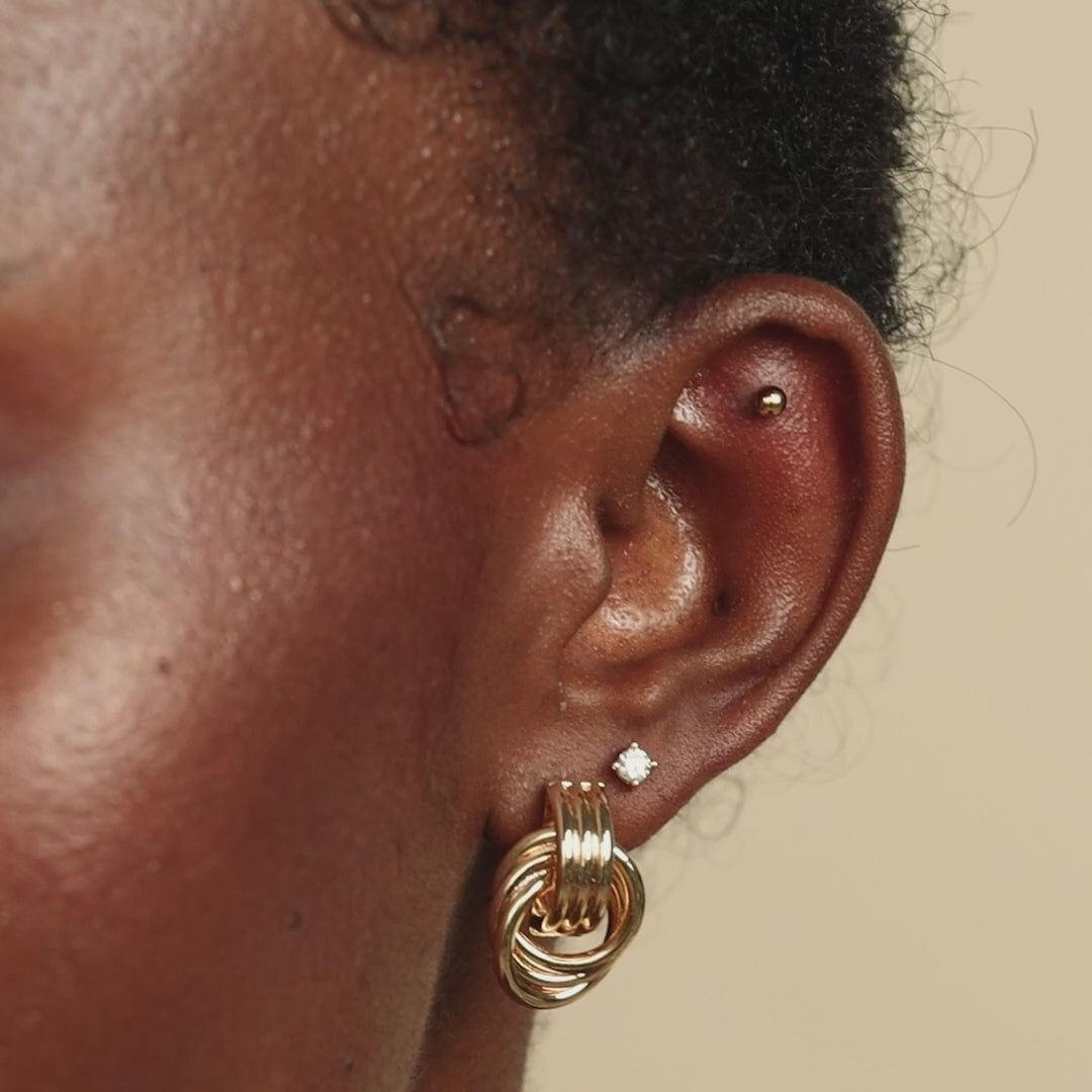 Side view of a person’s ear wearing the Mini Triple Knot Earring along with a small diamond stud and a gold cartilage piercing.