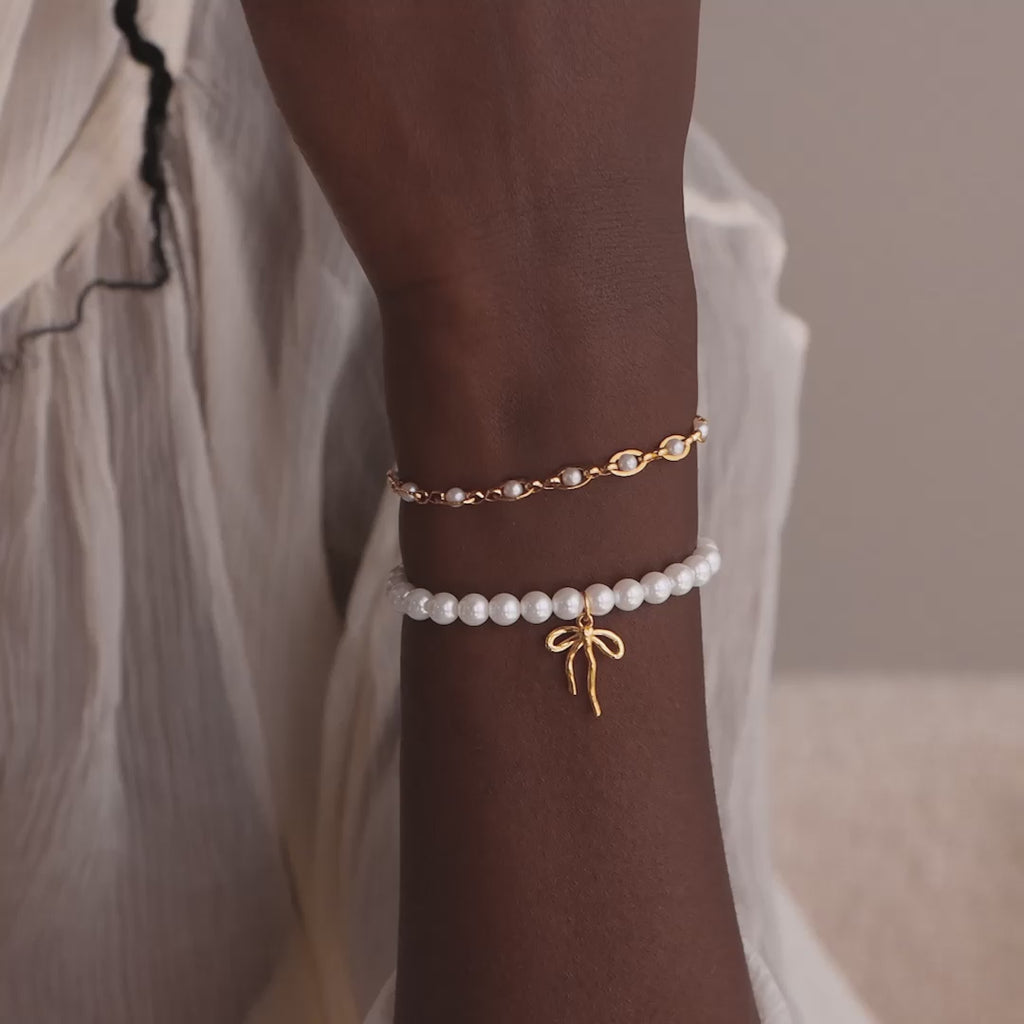 Model wearing a beaded pearl bracelet with a dainty gold ribbon bow charm, delicately stacked with a gold pearl eternity bracelet.