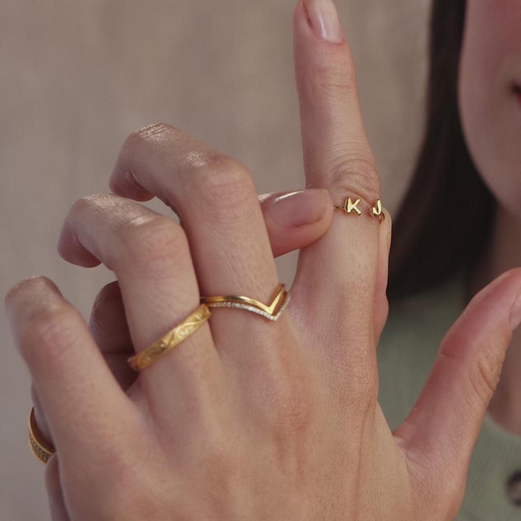 Detailed shot of stacked gold rings on hand, featuring a custom double initial ring with bubble letters K and J and a mix of textured and chevron bands.