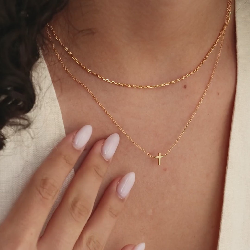 Model showcasing a dainty gold cross charm necklace, layered with a gold mariner chain necklace.