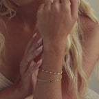 Close-up of model wearing two gold bracelets – a dainty Tiny Link Heart Bracelet and a pearl station bracelet stacked together.